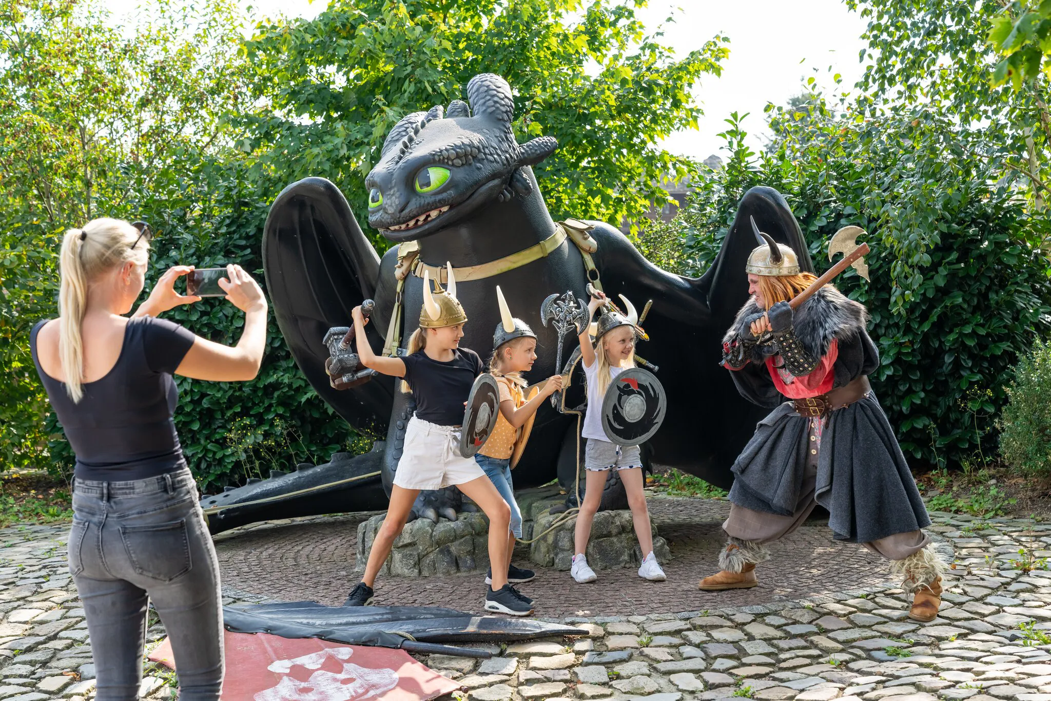 Eine Frau macht ein Foto von Ohnezahn , einem Wikinger und drei Mädchen mit Wikinger Utensilien im Heide Park Resort.