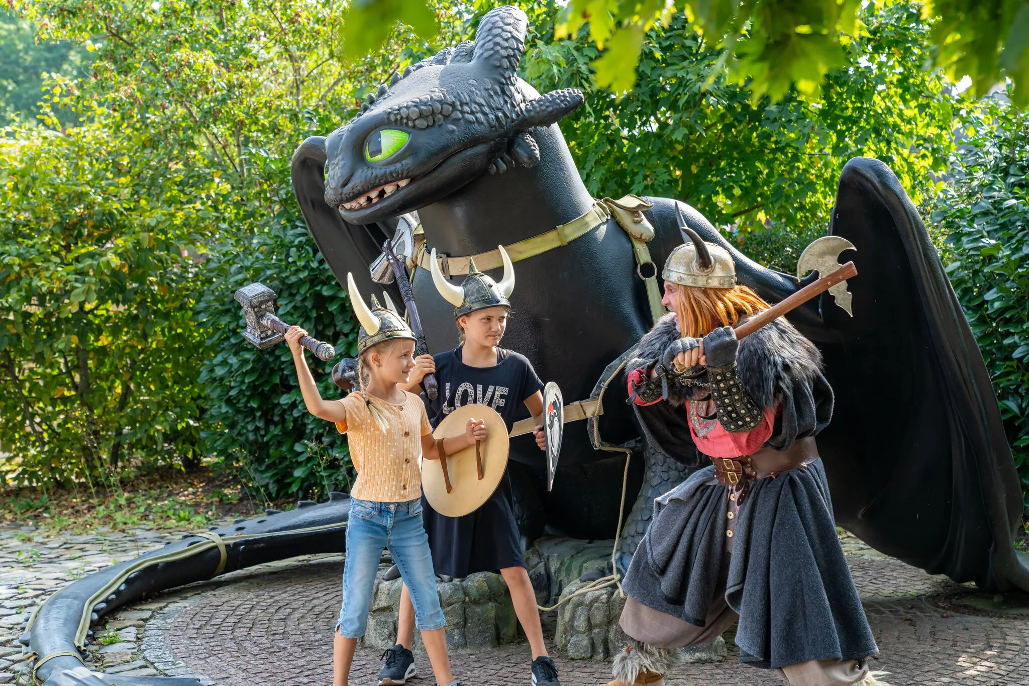 Ein Wikinger steht mit zwei Mädchen mit Wikinger-Utensilien vor einer großen Ohnezahn-Drachen-Figur